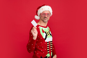 Fototapeta na wymiar Man with party glasses in holiday sweater and Santa hat on studio background. Santa man hold credit card. Christmas sale or special winter offer.