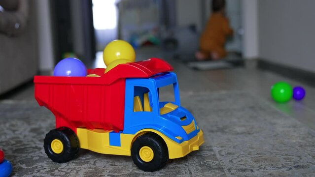 Beautiful bright toy lorry with plastic balls in the basket. Little baby crawls by the floor at backdrop in blur.