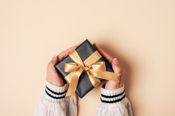 Girl's hands in white sweater holding black gift box with golden bow on beige background....