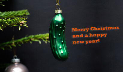 Christmas tree decorations. A green glass cucumber is hanging on a christmas tree. Green branches and a black background are visible.
