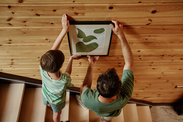 Top view of father and son hanging picture together on wooden wall in new house