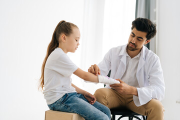 Professional male physician in uniform consulting injured little patient with broken hand wrapped in plaster bandage at checkup meeting on white background. Concept of child insurance and healthcare. - obrazy, fototapety, plakaty