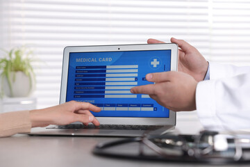 Doctor and patient using laptop to fill out medical card at table in clinic, closeup