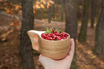Woman holding wooden cup with tasty lingonberries in forest, closeup