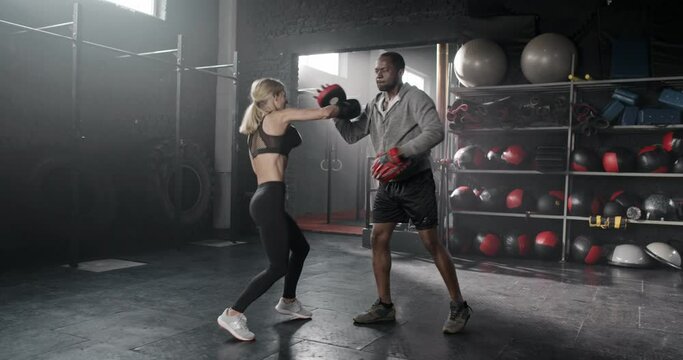 Beautiful blonde young woman training boxing punching and evading with personal trainer African experienced man. Professional strong coach with boxing paws teaching attractive female in boxing gloves.