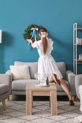 Young woman hanging Christmas wreath on blue wall in living room