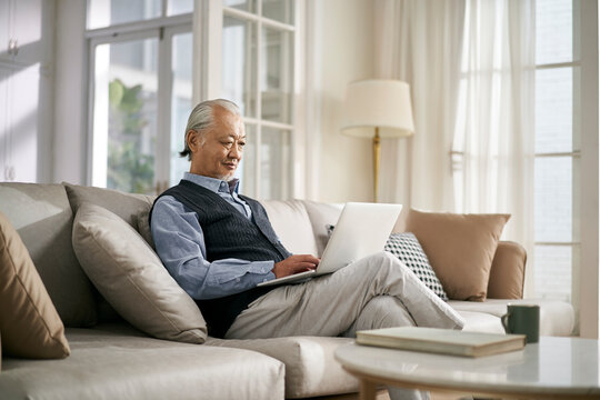 Senior Asian Man Sitting On Couch Looking At Notebook Computer