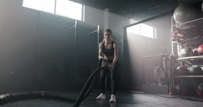 Attractive sporty fit young woman working out with battle rope doing crossfit exercise in modern gym. Beautiful focused sportswoman female wearing leggings and top training muscles.