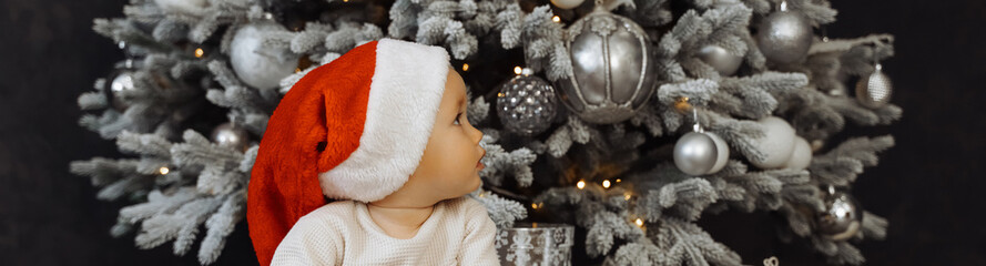 Portrait of newborn baby in Santa clothes and christmas hat, sits on the floor, winter snow landscape. banner