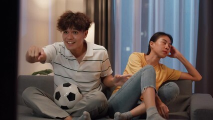 Asian new marriage couple sit on sofa husband excited cheering watching football sport game on television in living room at home. Young woman getting bored her partner watch online soccer on TV. - Powered by Adobe