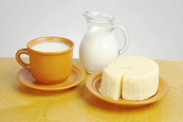 A cup of milk, a jug of milk and white cheese on a saucer.