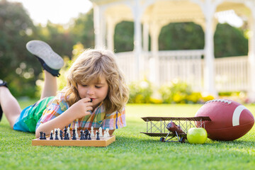 Summer games and outdoor activities for kids. Child playing chess game in spring backyard, laying on grass. Concentrated kid play chess. Kid playing board game outdoor. - obrazy, fototapety, plakaty