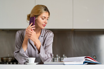 Fototapeta premium Young woman chatting on mobile phone, reading book or magazine and drink coffee on kitchen at home. Housewife mood in the morning.