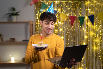 Virtual celebration birthday. Happy guy in party hat celebrating birthday online, using laptop for video call with friends and family on webcam, holding cake, standing in the living room