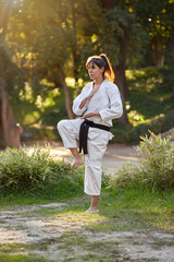 Woman in kimono and black belt standing in pose while practicing karate outdoors in nature. Sports...