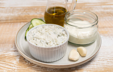 Obraz na płótnie Canvas Traditional greek yogurt sauce Tzatziki,dressing in white bowl, made of grated cucumbers, sour cream yogurt,olive oil,garlic,herbs with ingredients on wooden table. Close up, horizontal