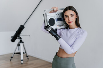 Attractive brunette hispanic girl in sportswear standing at home holds old fashion audio recorder looks aside. Young fit woman with artificial hand happy to be active. Orthopaedics and hi tech.