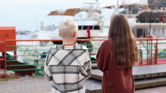 Back view of two friends girl and boy hanging out on vacation, children talking and having fun looking at sea and yachts