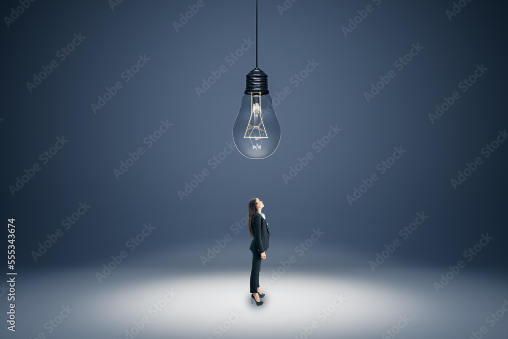 Poster Creative idea and brainstorming concept with businesswoman in black suit looking up at the ceiling with a big hanging light bulb on abstract empty grey area background