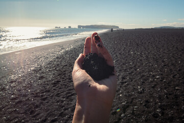 Close up female hand with sand concept photo. Seaside beauty. First person view photography with black basalt beach on background. High quality picture for wallpaper, travel blog, magazine, article
