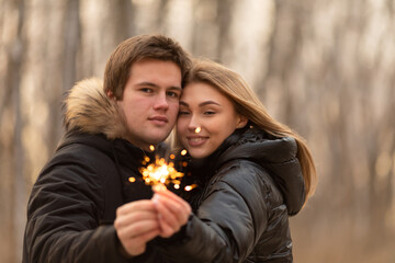 Obraz premium A loving couple in warm jackets kisses holding sparklers in their hands. Meet the winter holidays in a snowy forest. Couple in love, romance, happy valentine's day