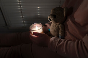A girl is in a dark room near the window and holds a candle and a teddy bear in her hands (close-up)