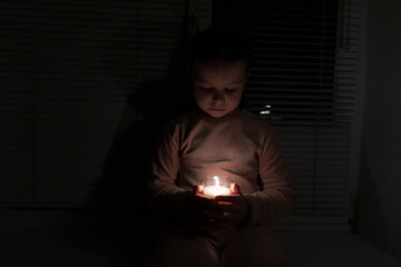 A girl in a dark room near the window holds a candle in a glass in her hands