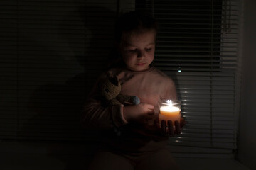 A girl in a dark room near the window holds a candle and a teddy bear in her hands