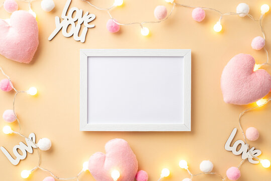 Valentine's Day concept. Top view photo of photo frame fluffy heart shaped toys inscriptions love you light bulb garland and soft pompons on isolated pastel beige background with empty space