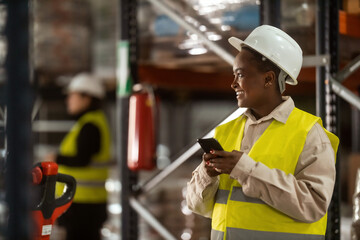 An African-American warehouse worker is using a mobile phone at work with a big smile on her face.