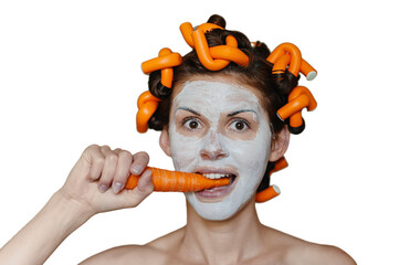 Young woman with cosmetic mask on her smiling face chewing fresh carrot, pretty housewife takes care of her skin with clay mask eating a fresh vegetable