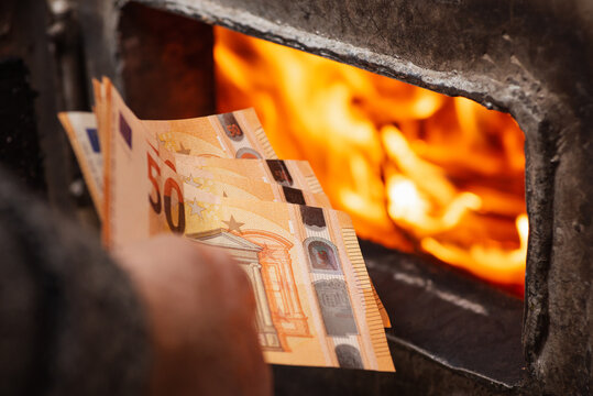 Euro bills in hand of a man near the heating boiler with opened doors and burning flames inside. Spending a lot of money to heat the house, expensive warming