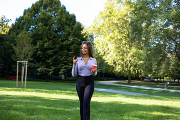 Fototapeta premium Woman walking in the park and reads a message on her smartphone. Portrait woman with brown hair, cup of coffee and mobile phone. Outdoor, public park, relaxed position and summer. 
