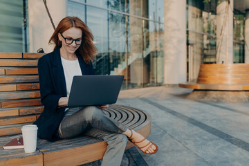 Beautiful businesswoman sitting on bench and working remotely on her project with laptop