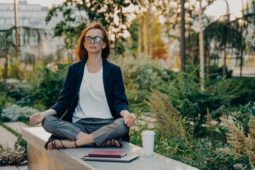 Full length shot of relaxed ginger woman meditates sits in lotus pose wears formal clothes