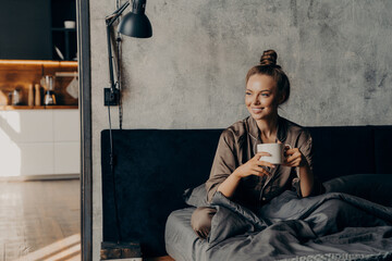 Joyful happy smiling caucasian female in satin cozy pajama holding cup of coffee