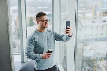 Smiling man in casual clothes standing near window with coffee and looking at smartphone in modern workspace with big window during daytime