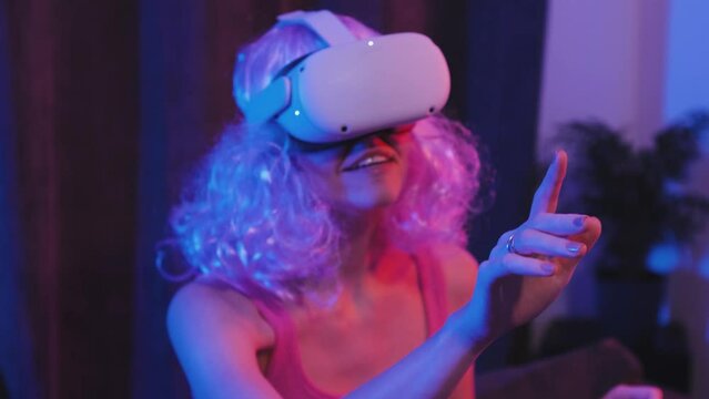 Young beautiful woman with pink hair uses VR headset for work or games in virtual reality. Girl gamer or streamer plays vr games. Pretty woman in vr headset in a room with neon lights