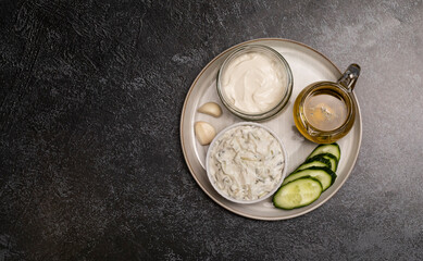 Traditional greek yogurt sauce Tzatziki,dressing in white bowl, made of grated cucumbers, sour cream yogurt,olive oil,garlic,herbs with ingredients on dark gray concrete table. Flatly, horizontal