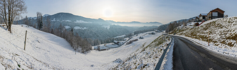 Frisch verschneite Bergstrasse, die sich ins Tal hinunter schlängelt, Bauernhaus am Strassenrand, Berge und Täler mit Wälder und Wiesen im Hintergrund, Abendstimmung im Winter, Hittisau mit Subersach,