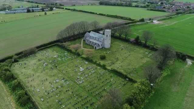 Aerial Drone Shot Flying Over Old Rural Medieval 11th Century Round Tower Church with Old Man Walking Through Graveyard in North Norfolk UK