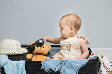 Happy baby girl sitting in travel suitcase puts sunglasses on a bear toy. Child with amused...