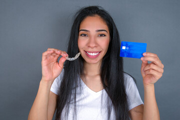 Young beautiful hispanic woman wearing white t-shirt over gray studio background, holding an invisible aligner on one hand and a credit card on the other hand. 