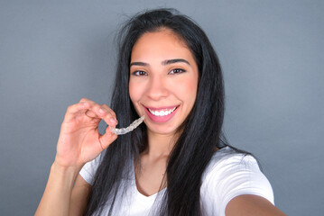 Young beautiful hispanic woman wearing white t-shirt over gray studio background, holding an invisible aligner on one hand and smiling.