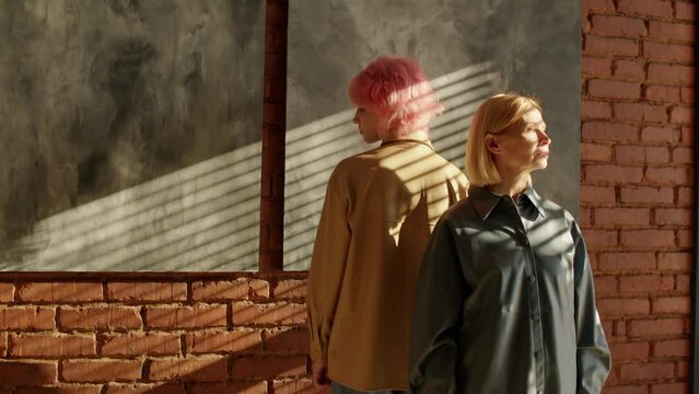 Conceptual portrait of modern young gen Z girl and mature woman wearing casual clothes standing back to back in loft room