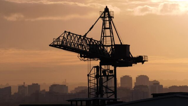 Silhouette of tall construction crane that turns arrow to lift delivery of building materials. Work on construction site. Against background of sunset and orange sunset in the city.