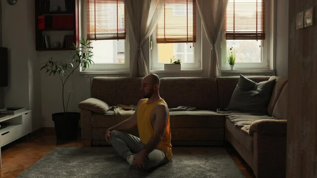 Meditation at home. Caucasian young man wearing sports clothes sits cross-legged on floor in living room and meditating. Sofa and windows at background. Concept of yoga, harmony and balance.