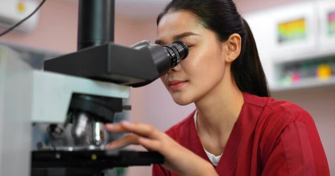 Close up shot, Asian young veterinarian female wearing uniform sitting and looking through a microscope, her hand slide sample of animals while working in lab,