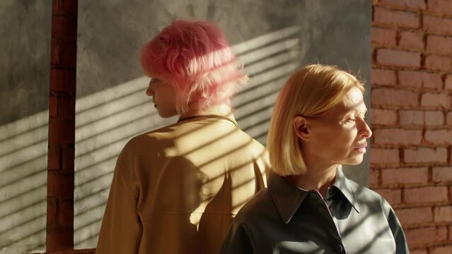 Studio portrait of young gen Z girl and mature gen X woman wearing casual clothes standing back to back in loft room looking away, generation gap concept