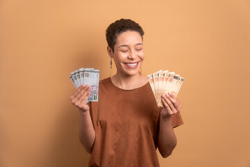 cheerful black business woman holding brazilian currency money in beige studio background. finance,...